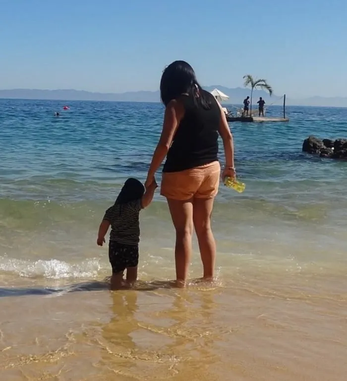 Kate paddling at the water's edge on a sunny beach, holding a little one's hand